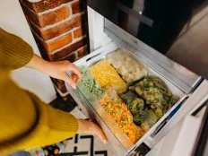 Woman putting container with frozen mixed vegetables to refrigerator