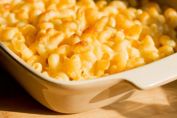 Homemade macaroni and cheese casserole sitting in the light of a window photograph.   The pasta has a cheesy buttery look and shows only the partial casserole dish. Macaroni and cheese is a comfort food for many.