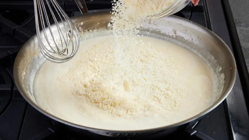 Photo showing  the sauce in the skillet made through step 5. Step 6 is starting: the heat is low so sauce is just warm, the Parmigiano-Reggiano is dumped in and a whisk is about to mix it for the Fettucini Alfredo recipe, as seen on Food Network.