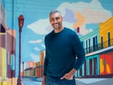 A man in a blue long sleeved shirt smiles while standing in front of a mural. 
