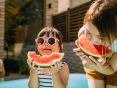 Adorable Asian little girl with sunglasses, enjoying summertime, eating fresh ripe watermelon with her mother. Summer vacation and family bonding time.